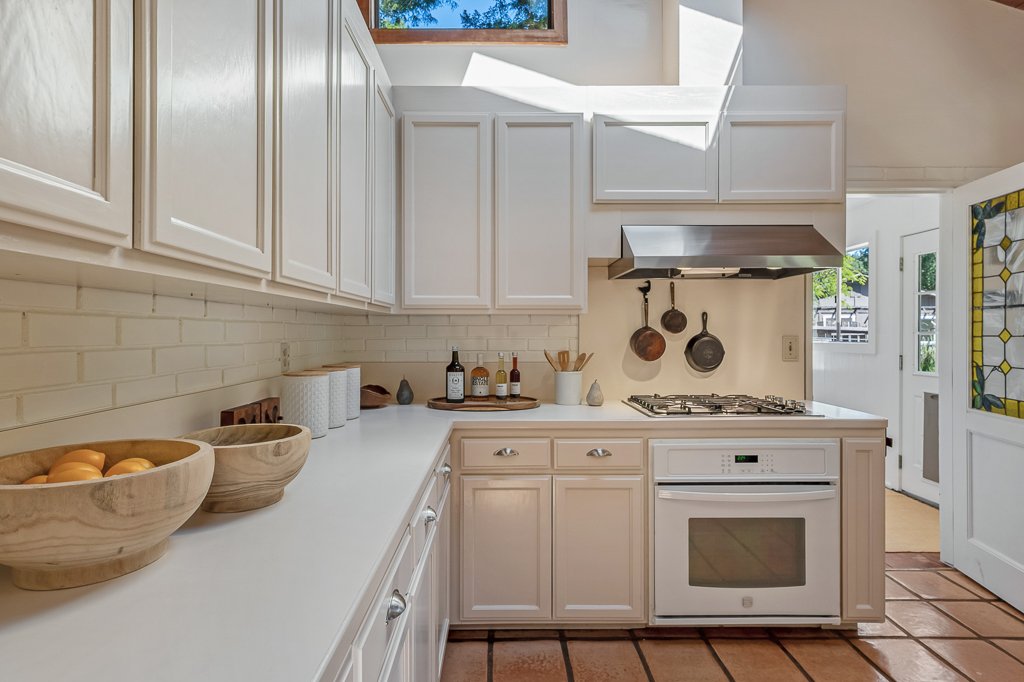Bright kitchen with white cabinets, beige tiled floor, and a white countertop. Two wooden bowls with fruit, bottles of oil or vinegar, and various kitchen tools are on the counter. Pots hang above the stove, and a window chimney is visible.