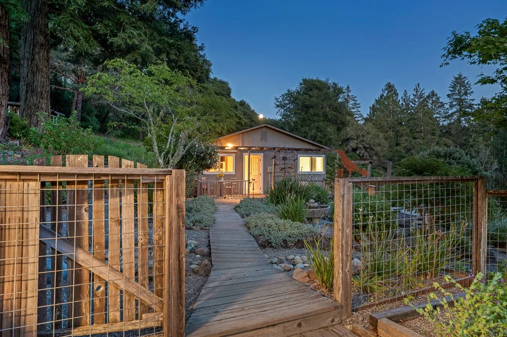 A cozy house surrounded by trees, with a wooden pathway leading to its front door, illuminated at dusk.
