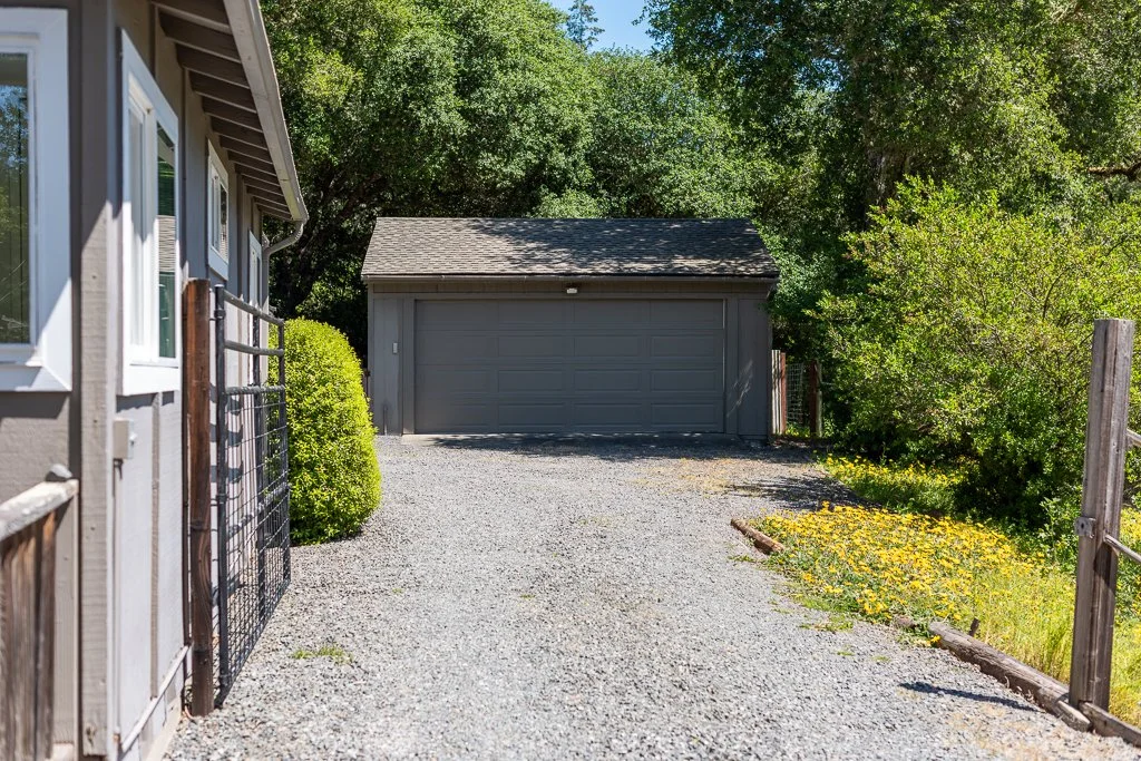A gravel driveway leading to a gray garage with trees and bushes on both sides.