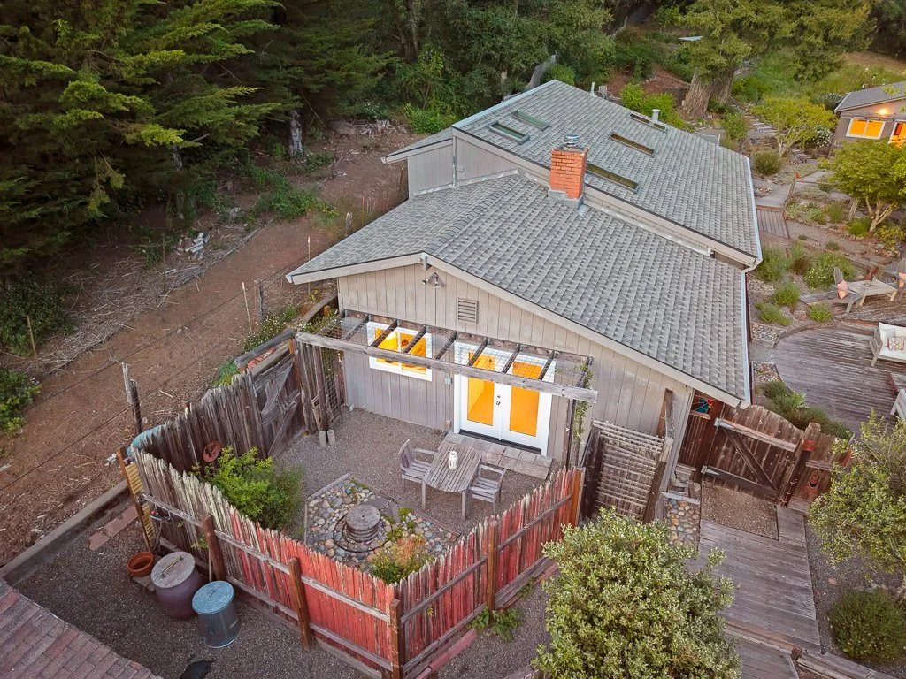 Aerial view of a house with a fenced backyard, patio, trees, and neighboring houses in a wooded area.