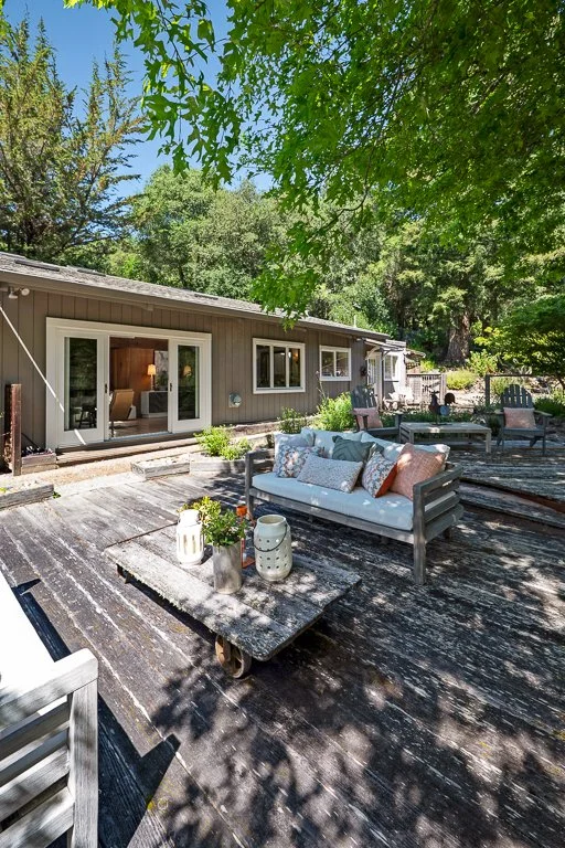 Outdoor patio area with rustic wooden furniture, cushions, and potted plants in front of a single-story house with sliding glass doors, surrounded by green trees.