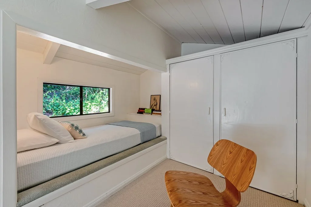 A cozy attic bedroom with a built-in window seat, white walls, and a small window showing green foliage outside. There is a bed with white bedding and a decorative pillow, a wooden chair, and white sliding closet doors.