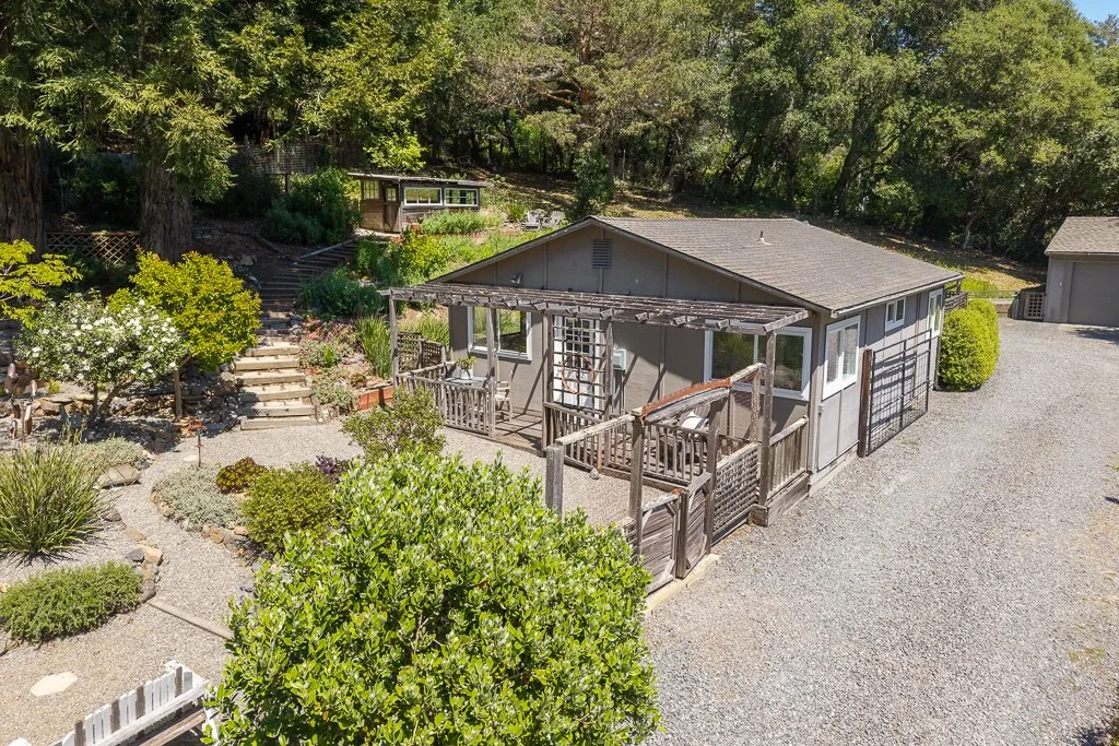 A backyard with a gray house, surrounded by trees and greenery, featuring a gravel pathway, a small wooden deck, and a set of stairs leading to a garden area with plants and flowers.