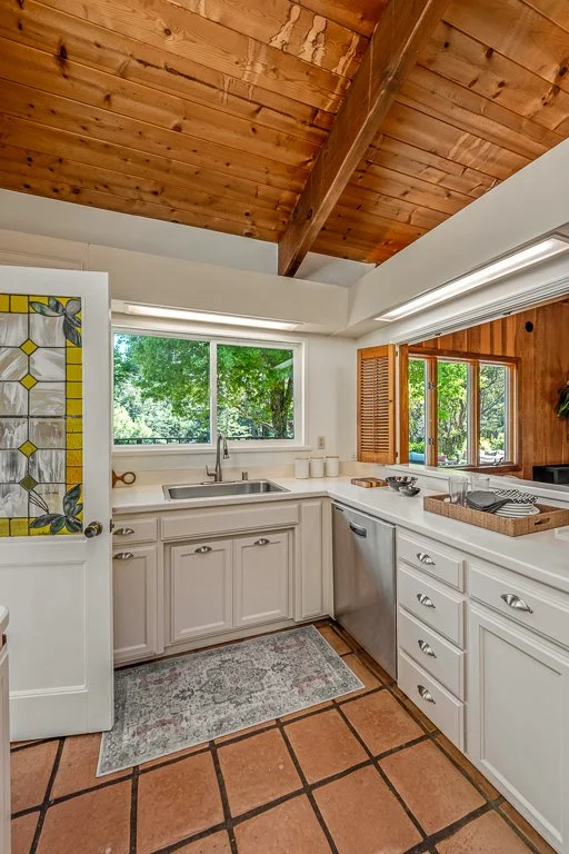 Kitchen with white cabinets, a sink under a large window, and a wooden ceiling. Outside greenery is visible through the windows.