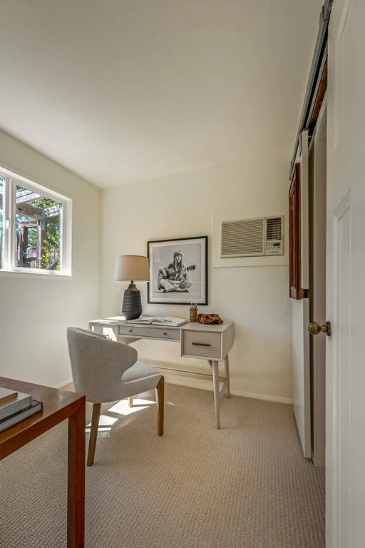 A small home office with a white desk, a gray upholstered chair, a black table lamp, a framed black and white photo on the wall, a window letting in daylight, and an air conditioning unit on the wall.