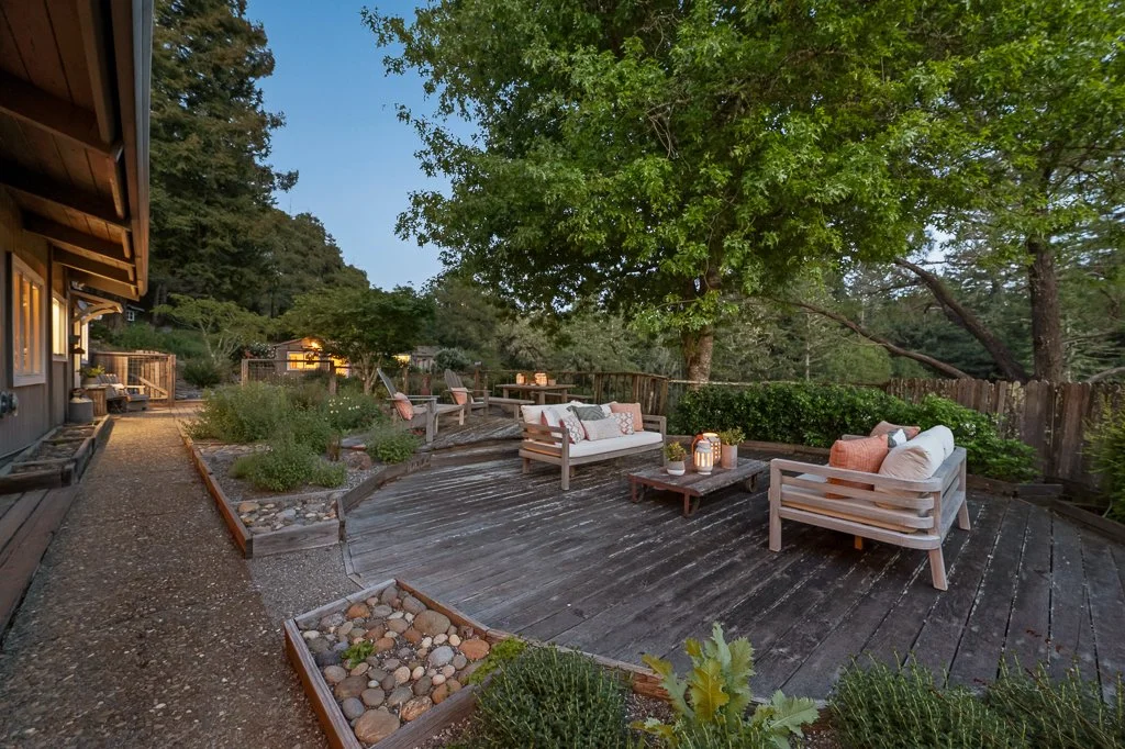 Outdoor wooden deck with seating area, including sofas with cushions, surrounded by trees and garden beds, in a backyard at dusk.