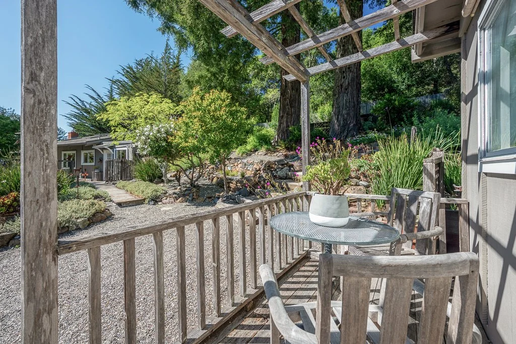 A cozy outdoor porch with a small round table, a potted plant, and wooden chairs, overlooking a garden with trees and shrubs.