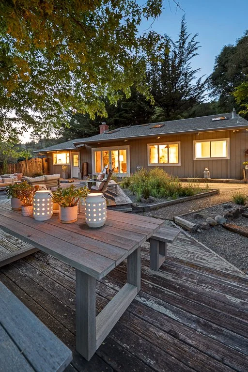 A backyard patio at dusk with lit lanterns on a wooden table, outdoor seating, and a house with illuminated windows surrounded by trees.