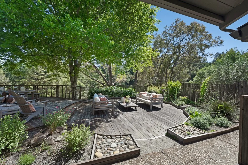 Outdoor wooden deck with seating and lush greenery, surrounded by trees and garden plants.