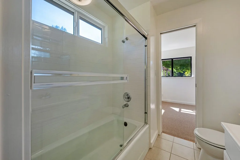 Bathroom with a glass-enclosed shower, white tiled walls, a small window, and a doorway leading to a room with a large window and carpeted floor.