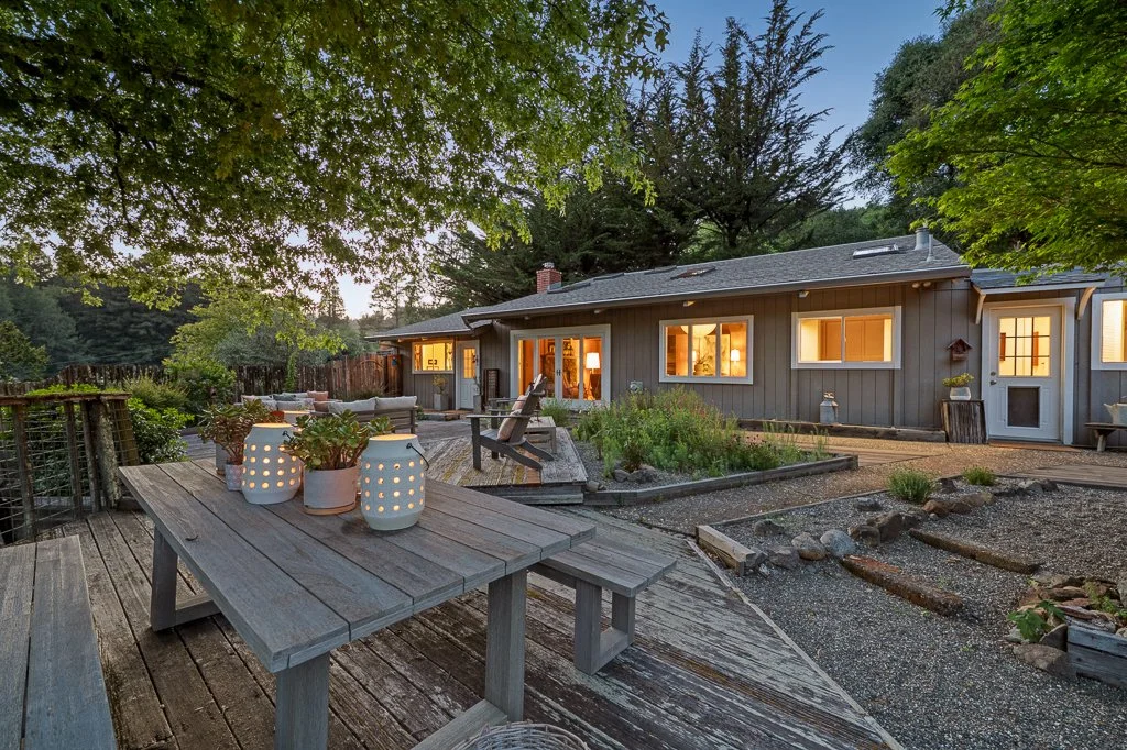 A cozy house with warm lights on, surrounded by a large backyard with trees, a wooden deck with outdoor furniture, and decorative lanterns in the late evening.