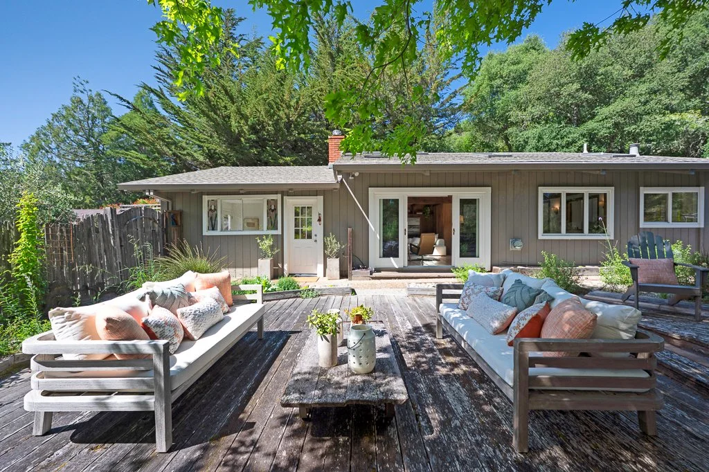 Outdoor wooden patio with two white sofas with pillows, a rustic coffee table, and an Adirondack chair, in front of a house with sliding glass doors and surrounded by green trees and plants.
