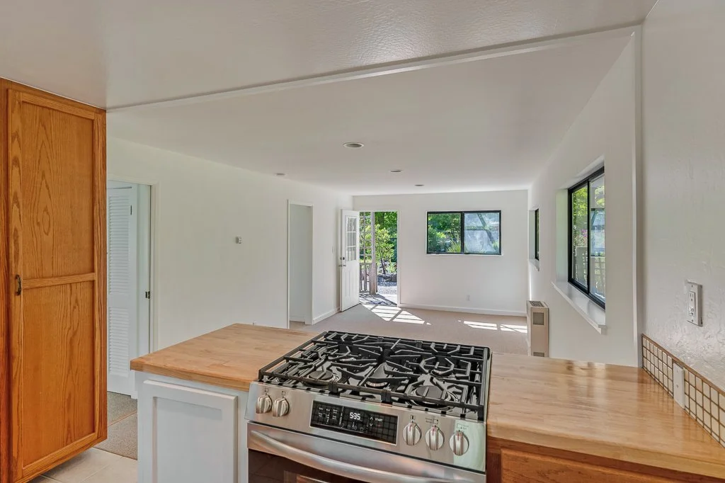 View from the kitchen showing a gas stove, wooden kitchen cabinets, and a bright living room with multiple windows and a door leading outside.