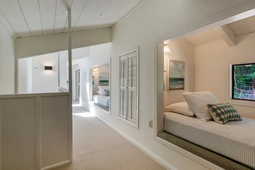 Interior view of a bright, cozy room with built-in beds, white walls, and a small window showing trees outside. There are framed paintings and checkered pillows on the beds.