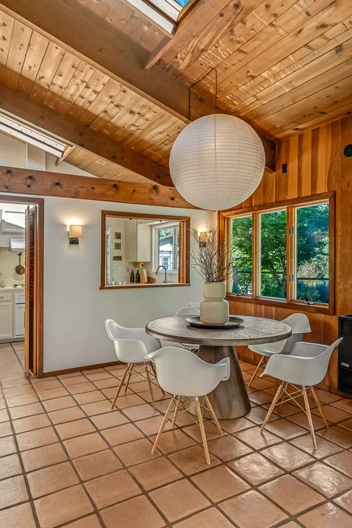Dining area with a round wooden table surrounded by six white modern chairs, a large white paper lantern hanging from a wooden vaulted ceiling, and a vase with dried branches as decoration, with windows showing green trees outside.