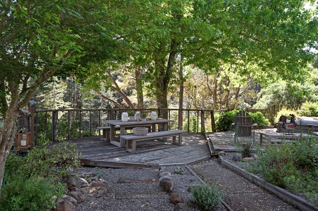 Wooden deck surrounded by trees and greenery, featuring a picnic table with four lanterns on top, a basket, seating area with chairs and a hot tub in a garden setting.