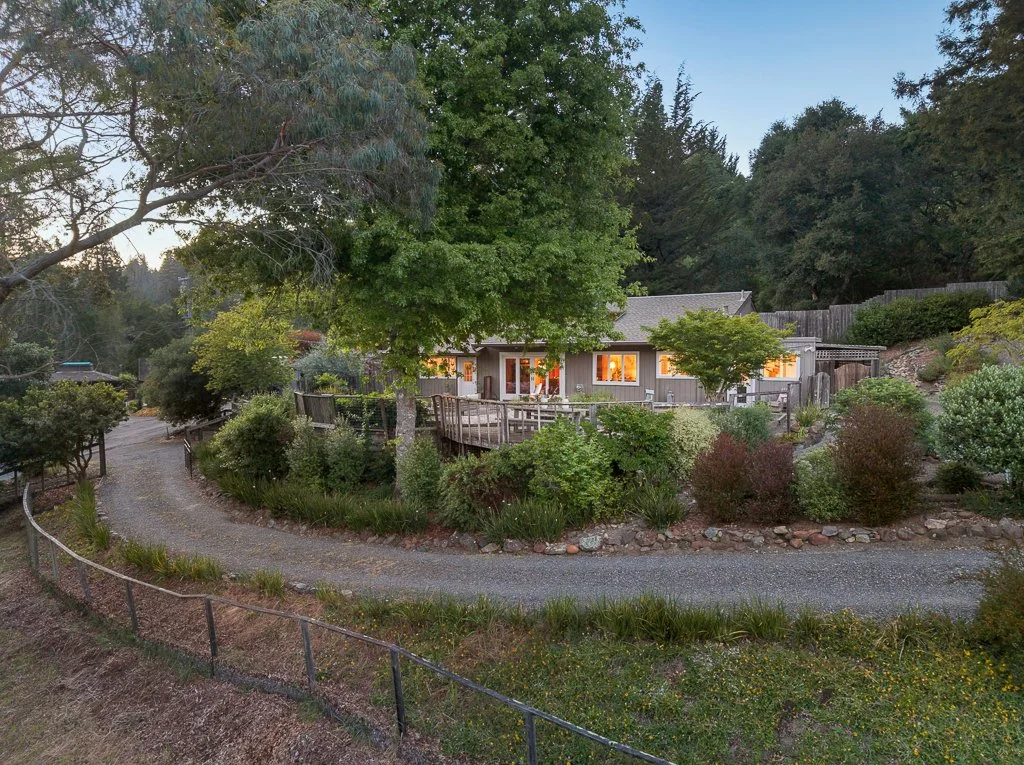 A house with glowing lights inside, surrounded by lush green trees and bushes, with a curved gravel driveway in front, set in a hilly, wooded area during dusk.