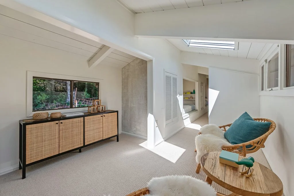 Bright bedroom with white walls, sloped ceiling, skylight, window, rattan chair with blue pillow, side table with decorative items, and built-in storage nook.
