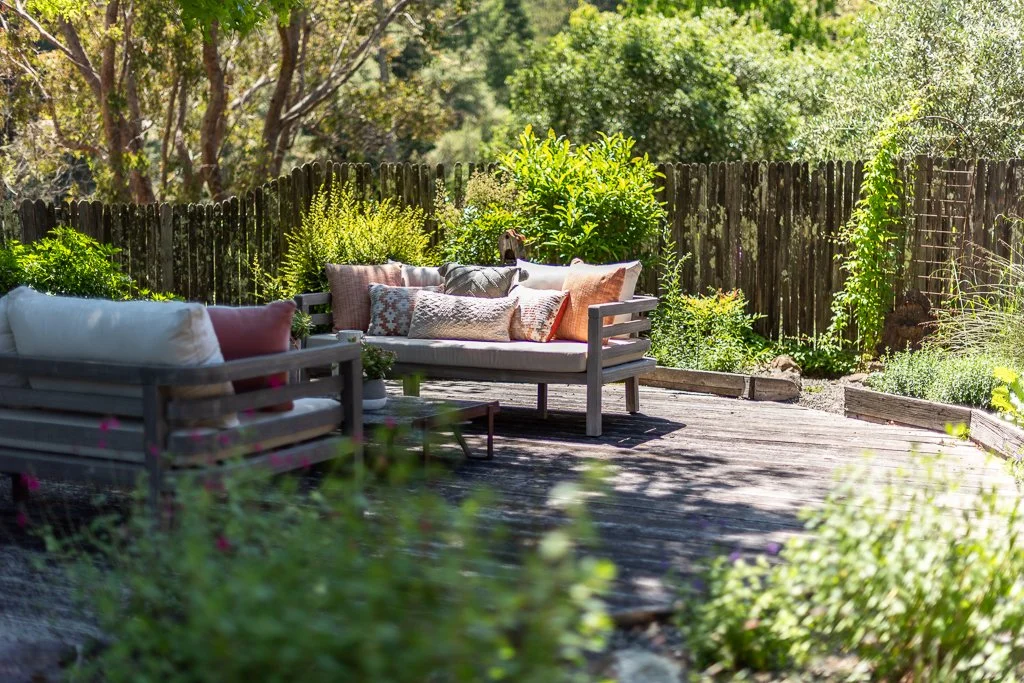 Outdoor wooden deck with lounge chairs and cushions, surrounded by lush garden greenery and a wooden fence.
