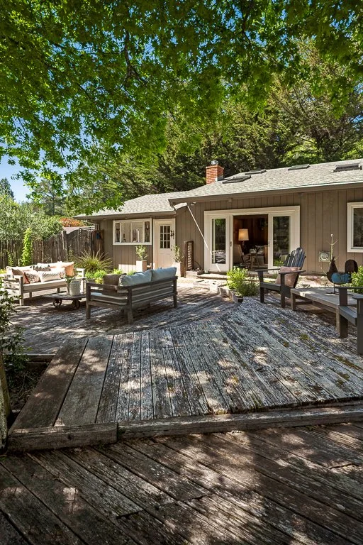 A backyard patio with outdoor seating, surrounded by green trees and a house with sliding door and windows.