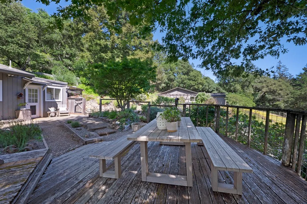 A wooden deck with a picnic table and benches, surrounded by a lush green garden with trees and plants, overlooking neighboring houses in a scenic outdoor setting.