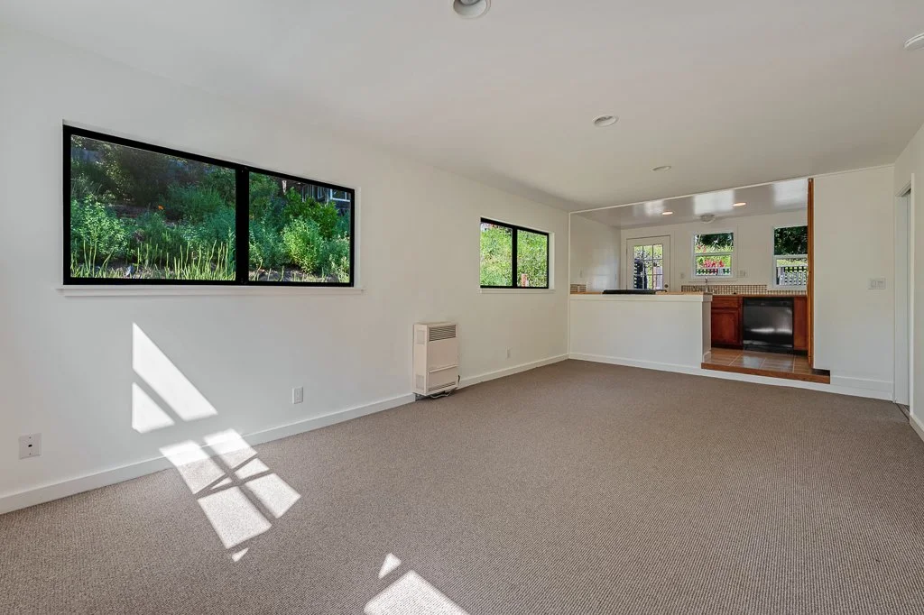 Empty living room with beige carpet, white walls, large window letting in sunlight, and view into the kitchen area with wooden cabinets.