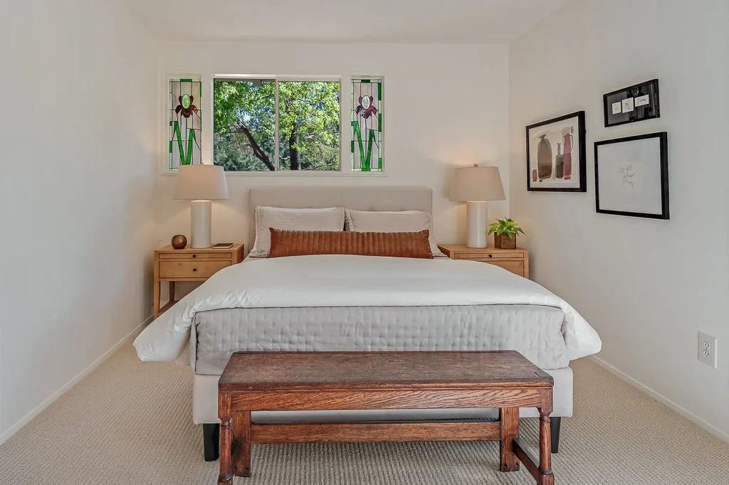 A bedroom with a white bed, wooden nightstands and lamps, framed art on the walls, and a window with colorful stained glass panels showing green trees outside.