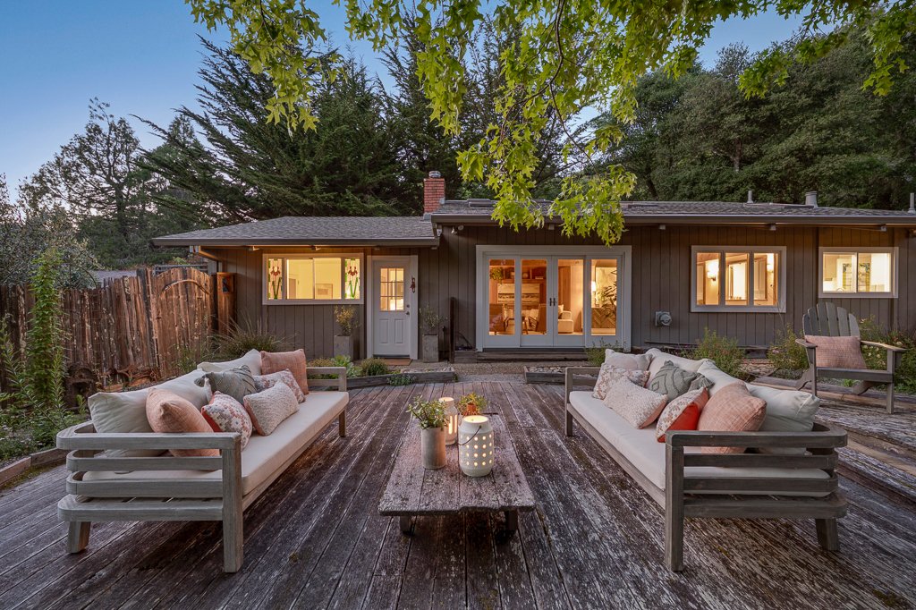 Outdoor patio with two white sofas and cushions, a wooden table with candles, adjacent to a dark gray house with lit windows and a sliding glass door, surrounded by trees and a weathered wooden fence.