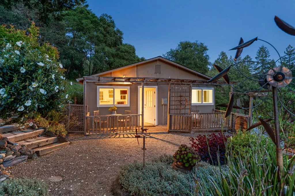 A small, cozy house with a wooden front porch, surrounded by a garden with various plants and flowers, illuminated at dusk.