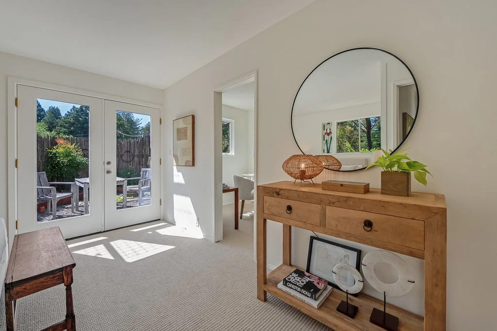A hallway with a glass door leading to an outdoor patio with chairs, an indoor wooden console table with decorative items, a round mirror, and a small window showing greenery outside.