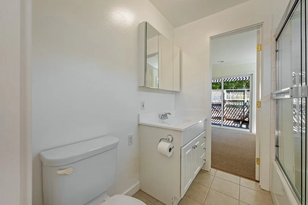 Bathroom with white toilet, vanity with mirror, small sink, and sliding glass door leading to an outdoor patio with wooden fence and outdoor furniture.