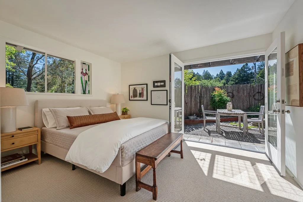 A bedroom with a large bed, nightstands with lamps, and a wooden bench, opening to a patio with a table, chairs, and greenery outside.