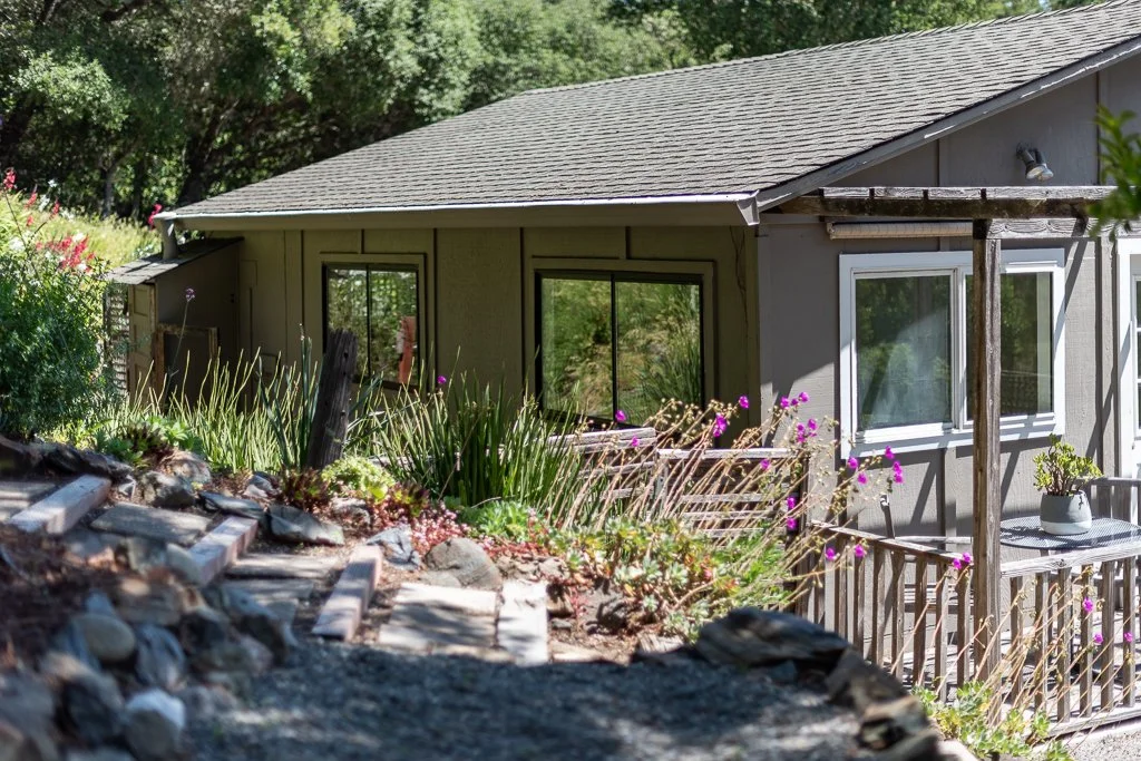 A house with a gray exterior, a sloped roof, and white-framed windows, surrounded by a garden with various plants and flowers, including pink blooms, and a stone pathway leading to a small porch with a potted plant.