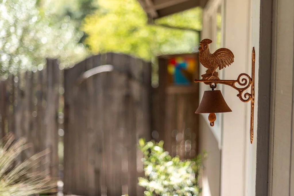 Close-up of a decorative wall-mounted rooster-shaped bell on the side of a house, with a blurred outdoor background of trees and a wooden fence.