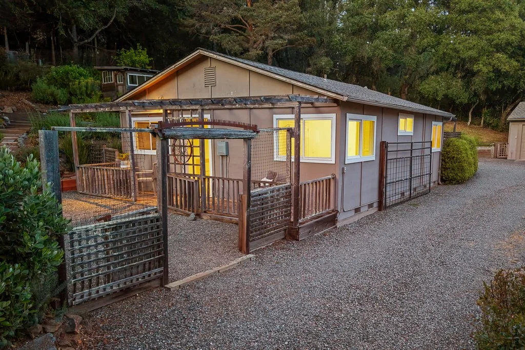 A small house with yellow-lit windows, a wooden deck, and a fenced yard, surrounded by trees and gravel ground.