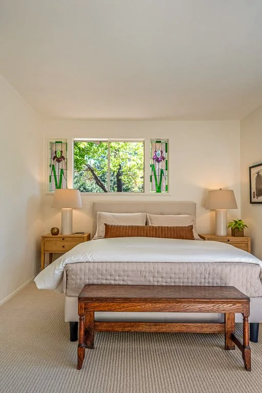 A bedroom with a bed, two nightstands with lamps, stained glass windows, and a wooden bench at the foot of the bed.