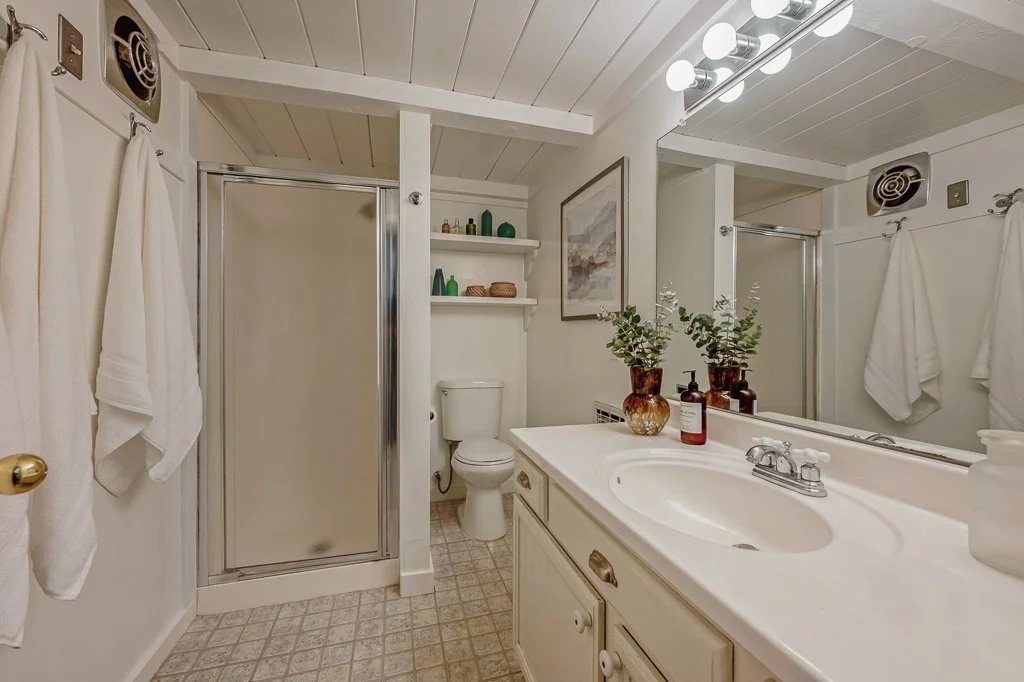 A small bathroom with a shower, toilet, and vanity with a sink. Decor includes plants, bottles, and framed artwork, with white towels hanging on the wall.