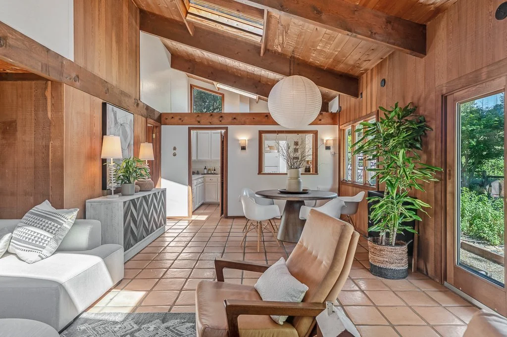 Living room with wooden walls and ceiling, terracotta tile floor, large windows, a white sofa with pillows, a beige armchair with a pillow, a round dining table with white chairs, a large potted plant, wall sconces, and a paper lantern pendant light.