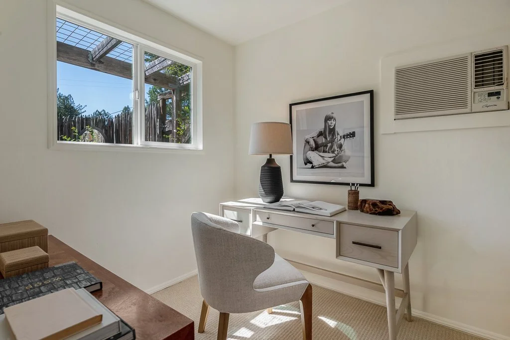 Small home office with a white desk, beige chair, black lamp, black and white framed photo of a girl with guitar, window with wooden fence outside, and air conditioning unit on a white wall.