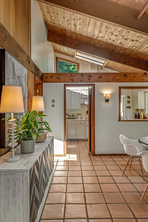 Interior view of a home showing a hallway leading to a kitchen with white cabinets, a dining area with white chairs and a round table, terracotta tiled flooring, and wooden ceiling beams with skylights.