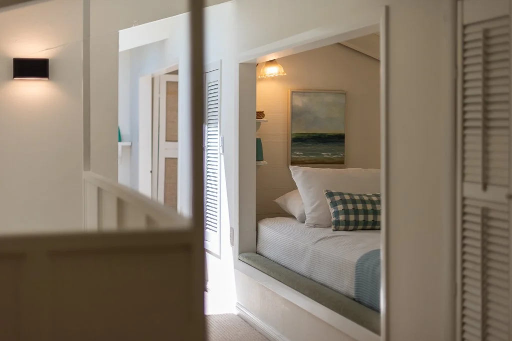 A cozy bedroom with white walls and a built-in window seat with cushions and pillows, framed by white walls and a door with shutter details, with painting and lamps on the walls.