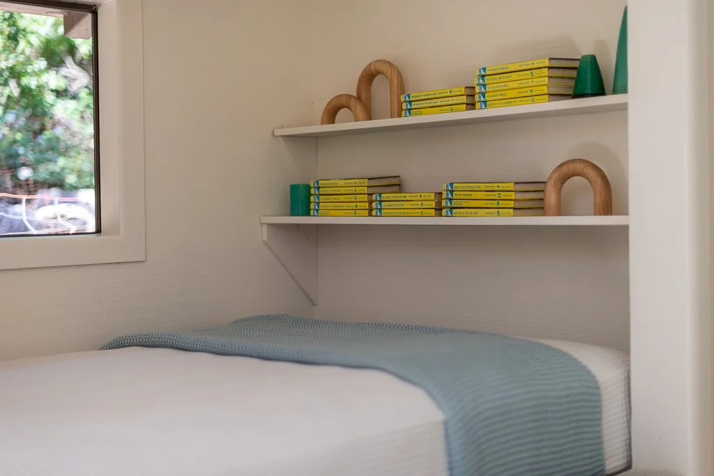 Bedroom with white bed and blue blanket, white wall-mounted shelves with yellow books, decorative wooden arches, green cone-shaped objects, and a window with trees outside.