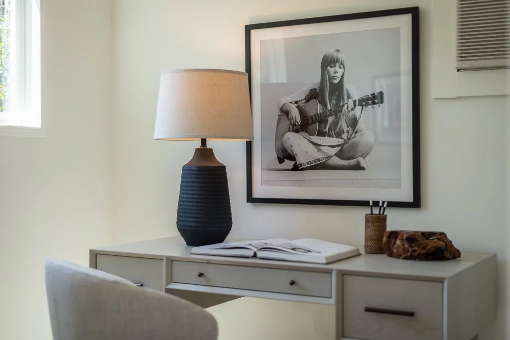 Close-up of a modern bedroom or study area with a white desk, a large black vase table lamp, an open book, and decorative items including a small container with pens and a wooden sculpture. Hanging on the wall above is a framed black-and-white photog