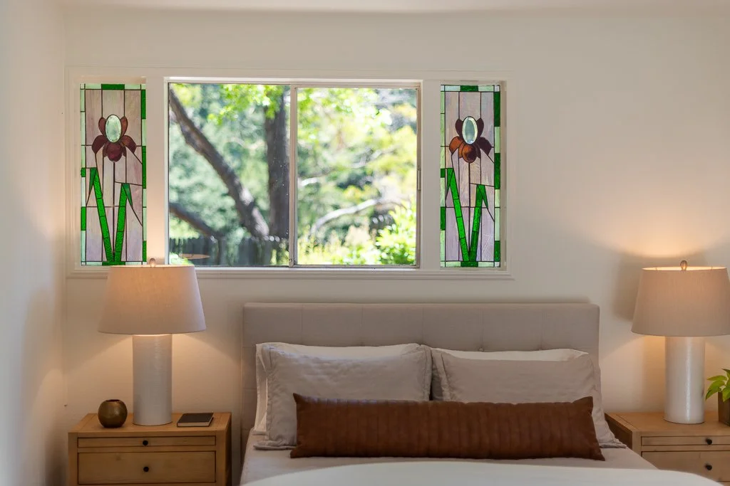 A bedroom with a large window featuring stained glass panels of flowers, a bed with white and brown pillows, two wooden nightstands with lamps, and a view of trees outside.
