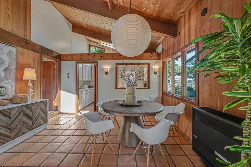 A cozy dining room with wooden walls and ceiling, a round wooden table with a large vase and twigs centerpiece, surrounded by white modern chairs, large windows on the right side, white cabinets in the adjacent kitchen, and warm lighting fixtures.
