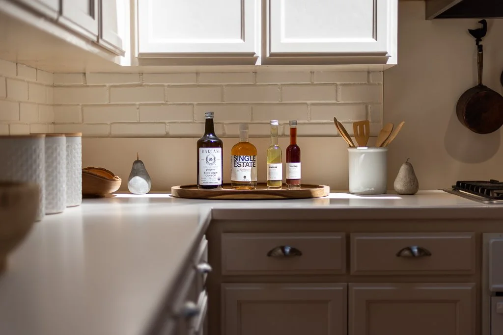 Kitchen counter with bottles of alcohol, wooden utensils in a container, decorative stones, and kitchen cabinets with a brick wall background.
