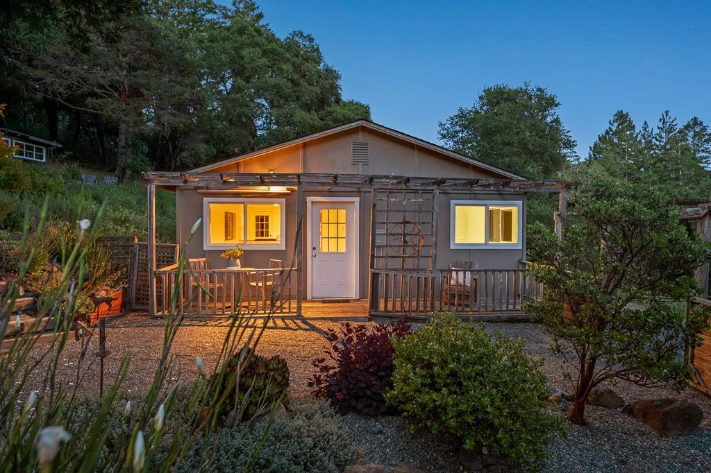 A small, rustic house at dusk with warm lights inside, surrounded by a garden with shrubs and trees.