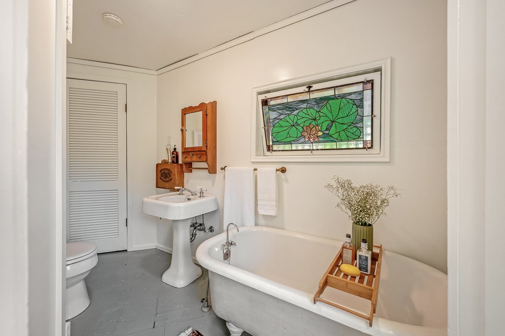 A vintage-style bathroom with a clawfoot bathtub, a small sink with a wooden cabinet above, a stained glass window, and a round toilet. Decor includes a tray with toiletries and a vase with flowers on the bathtub.
