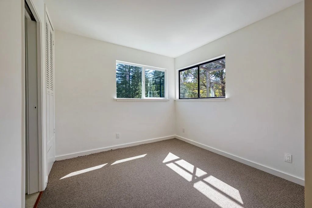 Empty room with white walls, two windows letting in natural light, beige carpet, and a closet with louvered doors.