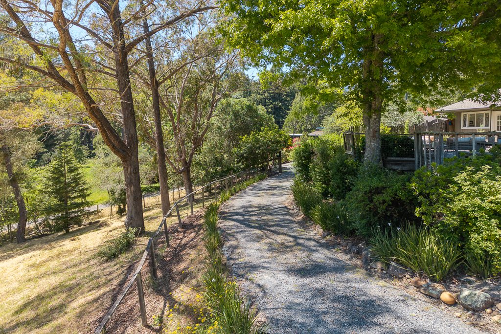 A gravel pathway winding through a lush, green garden with trees and bushes on both sides, and a house with a porch visible on the right.